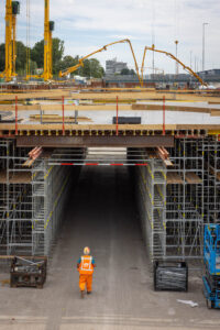 De bouwplaats van vooraf. Boven zijn diverse kranen bezig. Onder loopt iemand onder de werkzaamheden door.