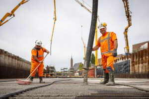 Twee personen die bezig zijn met het storten van beton. Links is de persoon aan het vegen. De persoon rechts kijkt in de camera terwijl hij zijn werk uitvoert.