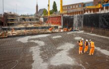 Een foto van afstand genomen van de bouwplaats. Met rechts een groep mensen bij het betonwerk en in de verte zijn veel stijgers te zien.