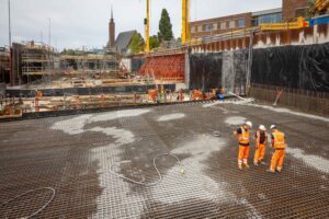 Een foto van afstand genomen van de bouwplaats. Met rechts een groep mensen bij het betonwerk en in de verte zijn veel stijgers te zien.