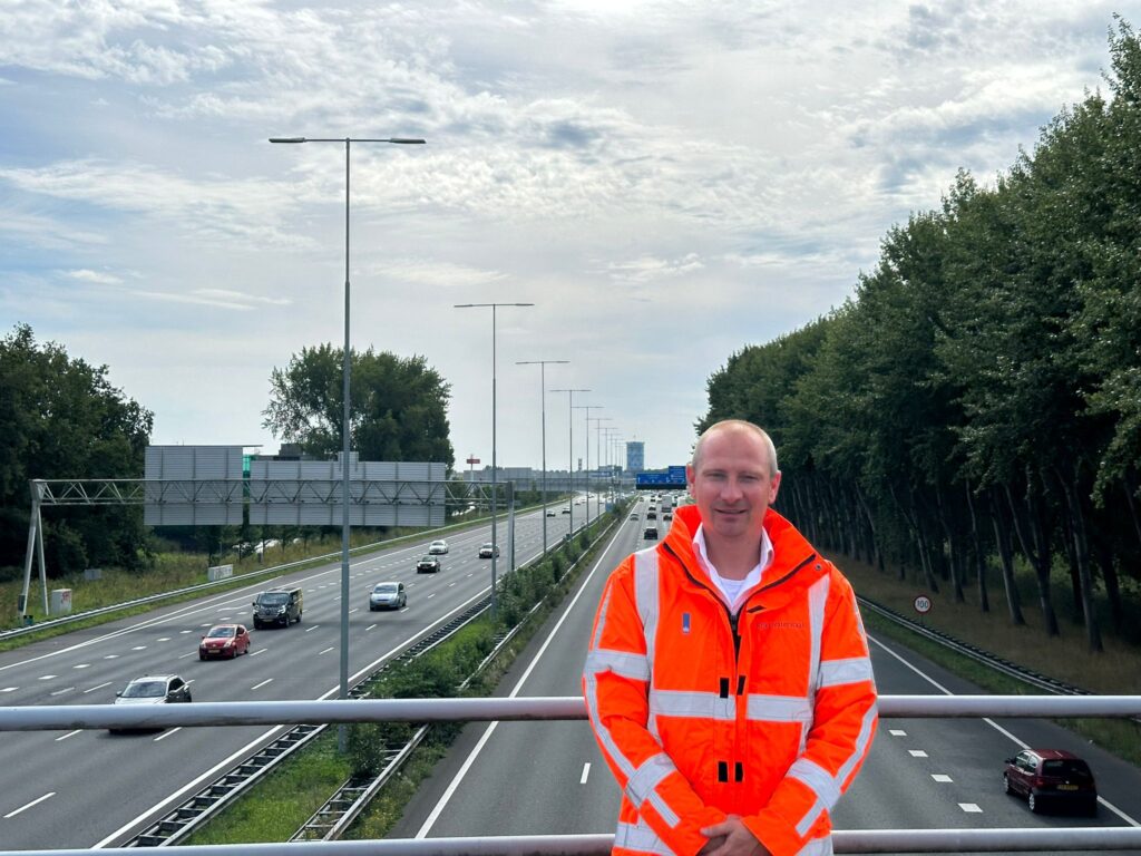 Verkeersmanager Bert Jan Zandhuis in een oranje jas op een viaduct over de A9, met de op de achtergrond de A9